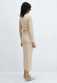 Beige, pitkähihainen mekko, jossa on vartalonmyötäinen siluetti, midi-pituus, teksturoitu kangas ja hillitty laskostus vyötäröllä. Litteät, hillityt sävyt kengät.