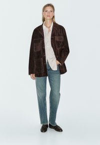 Giacca in suede marrone con ampie tasche, chiusura a bottoni, abbinata a una camicia a righe e jeans blu. Il modello è in piedi contro uno sfondo neutro.