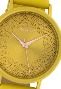 Montre en silicone jaune avec un cadran texturé, des aiguilles des heures et des minutes métalliques, et un design minimaliste. Logo "OOZOO" au-dessus du cadran.
