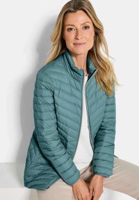 Woman with long blonde hair wearing a teal quilted jacket over a white top sitting against a white background.