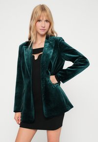 Βελούδινο πράσινο σακάκι με κομψά γραμμή, διακοσμημένο με διάσπαρτους ρόδινους κρυστάλλους, με καπακια και πλευρικές τσέπες. Μακριά μανίκια με μαύρες μανσέτες.