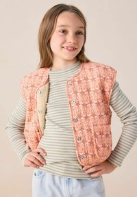 Gilet matelassé en orange clair avec des motifs floraux, doté de boutons-pression en argent et d'un intérieur en polaire, porté par-dessus un haut rayé.