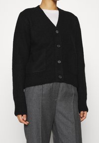 Černý vlněný cardigan s výstřihem do V, pěti knoflíky a dvěma předními kapsami. Žebrované manžety a lem, jemná textura. Nasažený přes šedé kalhoty.