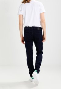 Jeans de mezclilla azul oscuro con corte ajustado, bolsillos traseros y una etiqueta de cuero en la cinturilla, combinados con una camiseta blanca y zapatillas blancas.