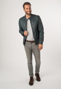 Giacca di pelle verde scuro con chiusura a zip e colletto a scatto, indossata sopra una t-shirt grigia, abbinata a jeans grigi e stivali marroni.