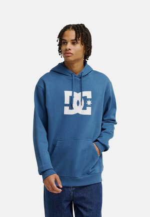 Jeune homme aux cheveux tressés portant un sweat à capuche bleu avec un logo blanc et un jean bleu, debout avec une main dans la poche.