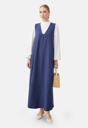 Donna che indossa un lungo vestito senza maniche blu navy sopra una maglia bianca a maniche lunghe, con una borsa intrecciata in mano, in piedi su uno sfondo bianco.