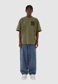 Oliven grøn oversized t-shirt med sort logo på brystet, parret med vide lyseblå denimjeans og hvide sneakers.