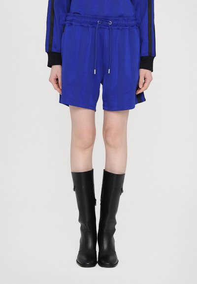 Des shorts bleu à cordon de serrage présentent un tissu texturé et des poches latérales, associés à des bottes noires en cuir montantes et une manche noire en accent.