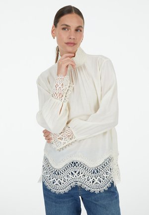 White blouse with a high neckline, featuring intricate lace detailing on sleeves and hem, long sleeves, and a relaxed fit.