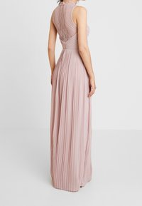 Lang, mouwloze blush jurk met een geplooide rok en ingewikkelde kantdetails aan de achterkant, met een aansluitend lijfje en gestructureerde stof.