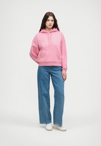 Rosa Kapuzenpullover mit Kängurutasche vorne, lockerem Schnitt und Kordelzug. Kombiniert mit weitgeschnittenen blauen Jeans und weißen Turnschuhen.