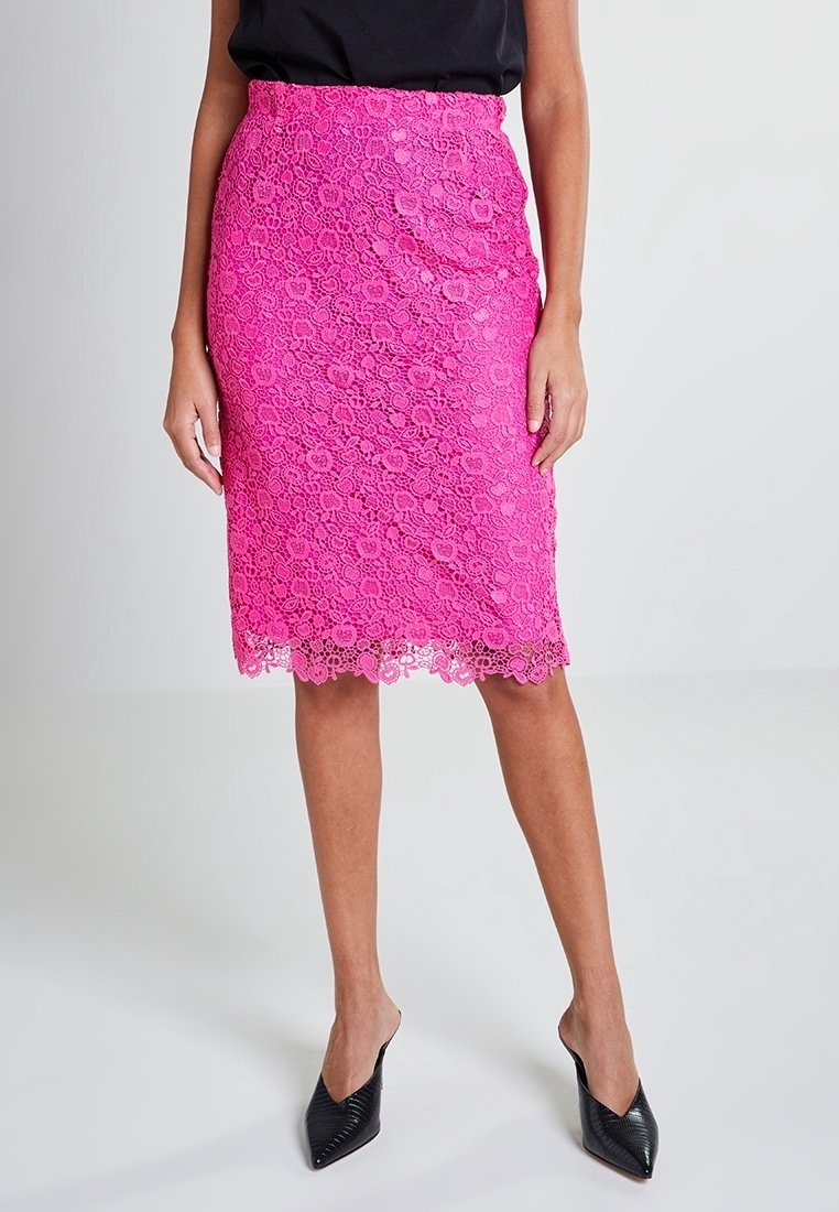 Žena v jasně růžové květinové krajkové sukni po kolena a černých lodičkách s špičatou špičkou, stojící na neutrálním pozadí.