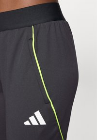 Svarte treningbukser med grønn piping på siden og et hvitt Adidas-logo. Har elastisk midje og en sidelomme.