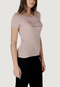 Camiseta de manga corta de color rosa claro con un diseño ajustado, que presenta un logo metálico "EA7 Emporio Armani" en dorado en la parte delantera.