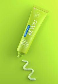 Grön tandkrämstub med böjd munstycke, märkt "Curaprox Be You," med detaljer av äppel och aloe, som dispenserar en ljusgrön pasta.