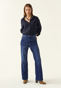 Jeans de tiro alto en azul oscuro combinados con una camisa de botones azul marino y botas de tacón marrones. El conjunto presenta un corte clásico y una textura suave.