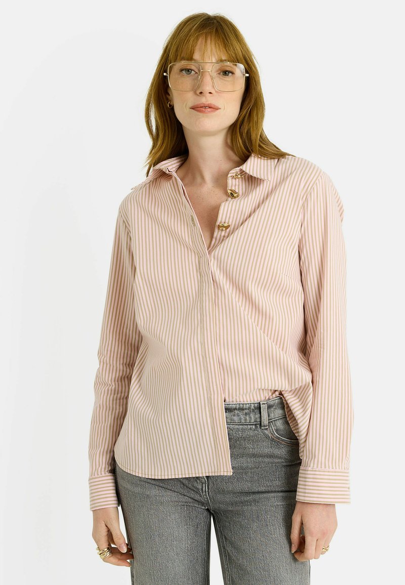 Camisa de botones rosa a rayas con mangas largas, con botones dorados, cuello y dobladillo curvado. Combinada con jeans grises.