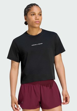 Ung kvinde med fletninger iført en sort "Adidas Tennis" t-shirt og vinrøde sportsshorts, poserer mod en ensfarvet baggrund.