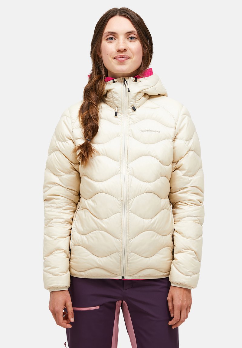 Doudoune beige avec motif en vagues, fermeture éclair à l'avant et capuche ajustable. Accentuée d'une doublure rose. Portée avec un pantalon violet.