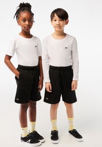 Czarne szorty z elastycznym pasem, w zestawie z białymi koszulami z długim rękawem z zielonym logo. Czarne sneakersy i skarpetki w żółte paski.