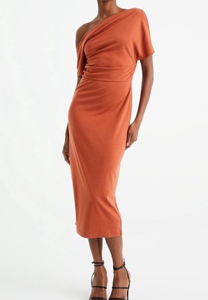 Nainen pukeutuneena ruosteenväriseen, yhdellä olkapäällä olevaan midi-mittaisen mekon, jossa on lyhyt hiha, ja mustat nilkkaremmi-korkokengät, seisomassa yksinkertaisen valkoisen taustan edessä.