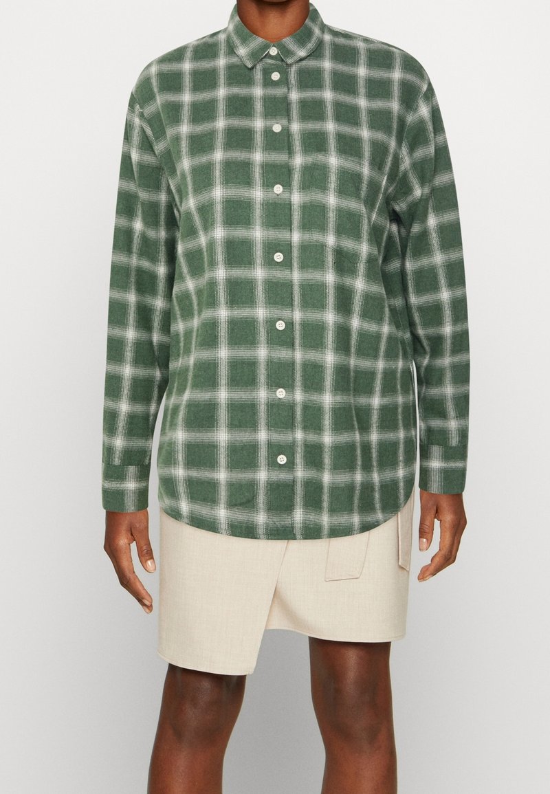 Personne portant une chemise à manches longues à carreaux verts et blancs boutonnée et une jupe asymétrique beige.