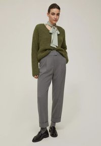 Maglione verde oliva con scollatura bassa, pantaloni grigi su misura con risvolti e mocassini neri con finitura testurizzata.