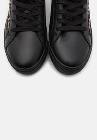 Černé kožené tenisky s texturovaným svrškem, kulatou špičkou, černými tkaničkami a zlatým zipem podél strany. Hladký povrch, nízký profil.