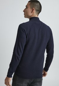 Námornícky sveter s dlhým rukávom z pleteniny, ktorý má vysoký golier, ribovanú textúru a priliehavý strih. Tkanina vyzerá mäkko a je mierne elastická.