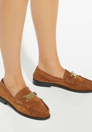 Voeten in bruine suède loafers met gouden kettingdetail en franjes, tegen een effen witte achtergrond.