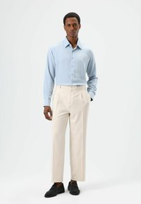 Light blue long-sleeve shirt with a pointed collar, paired with cream pleated trousers and black suede shoes. Flat texture, tailored fit.