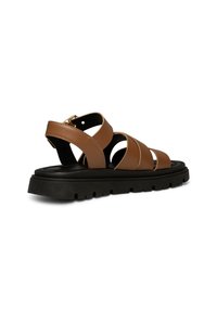 Hnědé kožené sandály s několika křížovými popruhy, černou gumovou podrážkou a plochou podrážkou. Na boku mají zlatý kovový detail.