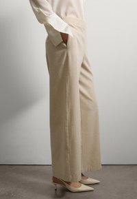 Personne portant un pantalon large beige, des escarpins ivoire à talons pointus, et une chemise à manches longues crème, avec la main dans la poche, se tenant à l'intérieur.