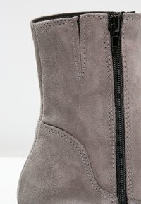 Bottine en daim gris avec fermeture éclair noire visible et couture détaillée sur fond blanc.
