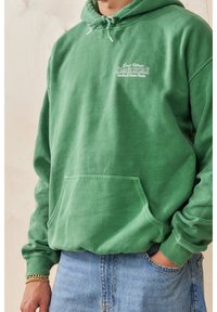 Zelená mikina s kapucí z měkké látky, vybavená střední kapsou, nastavitelnou stahovací šňůrkou na kapuci a bílým vyšitým nápisem na hrudi.