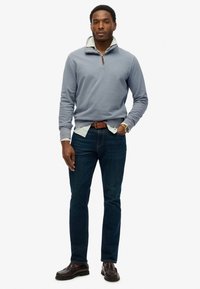 Lichtblauwe pullover met een rits en gestreepte kraag, gecombineerd met donkere jeans, een bruine riem en donkerbruine instappers.