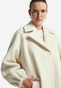 Vit ullblandad kappa med bred krage, avslappnad passform och rynkade manschetter. Framfickorna och den släta texturen förbättrar dess design.