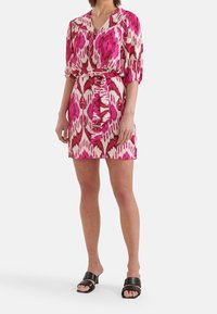 Kortärmad klänning i rosa och krämfärg med ett abstrakt mönster, med V-ringning och knytning vid midjan. Bärs med svarta sandaler.