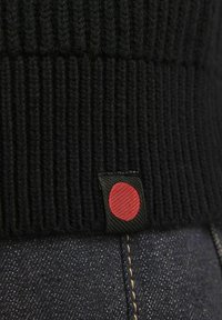 Schwarzer gerippter Strickstoff mit einem roten Logotag auf einem dunklen Denim-Hintergrund, der Textur und ein sauberes Design präsentiert.