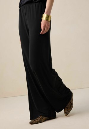 Schwarze, fließende Hosen aus leichtem Stoff, kombiniert mit spitzen Schuhen im Schlangenmuster. Ein goldener Armreif ziert eine Hand.