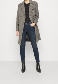 Abrigo de cuadros gris de doble botonadura, camiseta negra, jeans skinny oscuros y botas de tobillo negras con tacón. Cuenta con un bolsillo lateral y tejido texturizado.