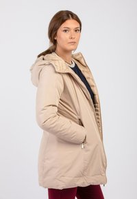 Beige geïsoleerde jas met een capuchon, voorzien van een ritssluiting aan de voorkant, zijzakken en een gevoerde binnenvoering. Soepele buitenoppervlakte.