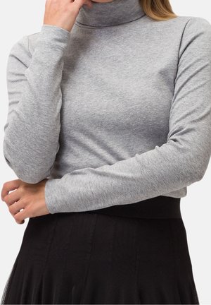 Femme portant un haut gris clair à manches longues et col roulé, les mains croisées sur une jupe noire.