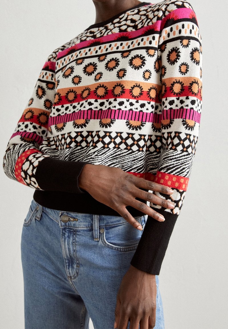 Personne portant un pull multicolore à motifs avec des poignets noirs et un jean bleu clair, croisant un bras sur l'autre.