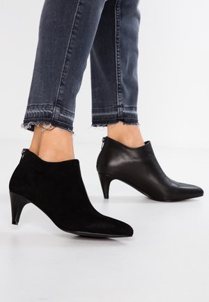 Woman wearing mismatched ankle boots; one black suede with a zipper back, the other black leather with a pointed toe and low heel.