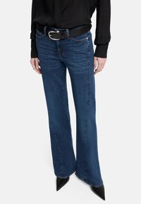 Blauwe denim flared jeans met een lichte vervaging, voorzien van vijf zakken en een zwarte leren riem met een zilveren gesp, gecombineerd met een zwarte top.