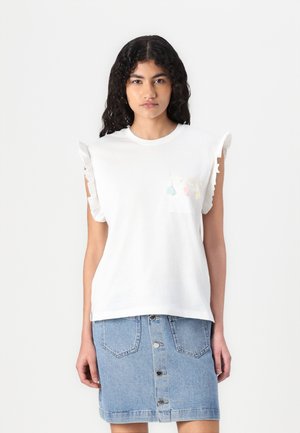 T-shirt blanc à manches courtes avec des épaules froncées et une poche avant ornée de pompons colorés. Associé à une jupe en denim bleu.