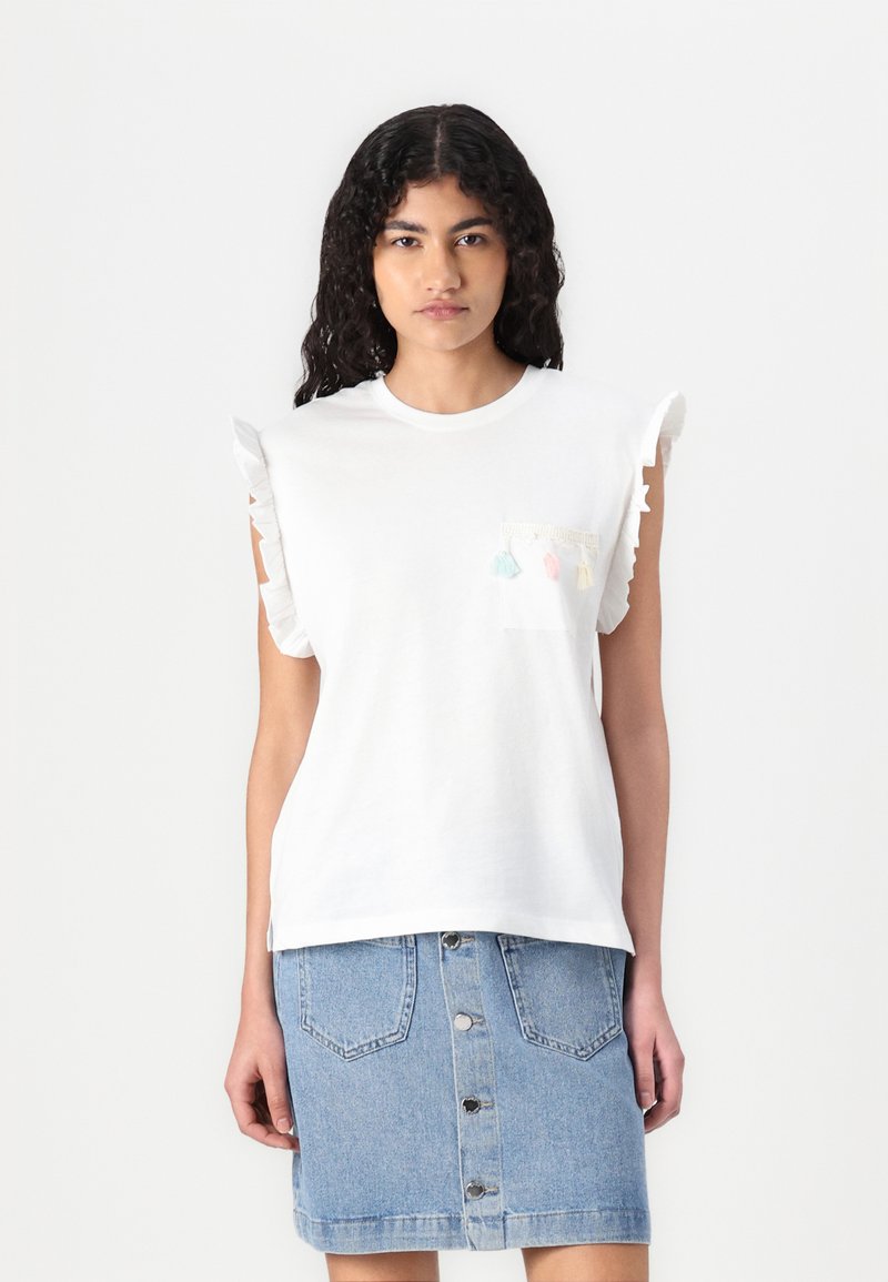 T-shirt blanc à manches courtes avec des épaules froncées et une poche avant ornée de pompons colorés. Associé à une jupe en denim bleu.