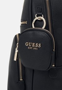 Zaino nero texturizzato con una tasca rotonda con zip e hardware dorato, che presenta il logo "GUESS EST 1981" in lettere dorate.